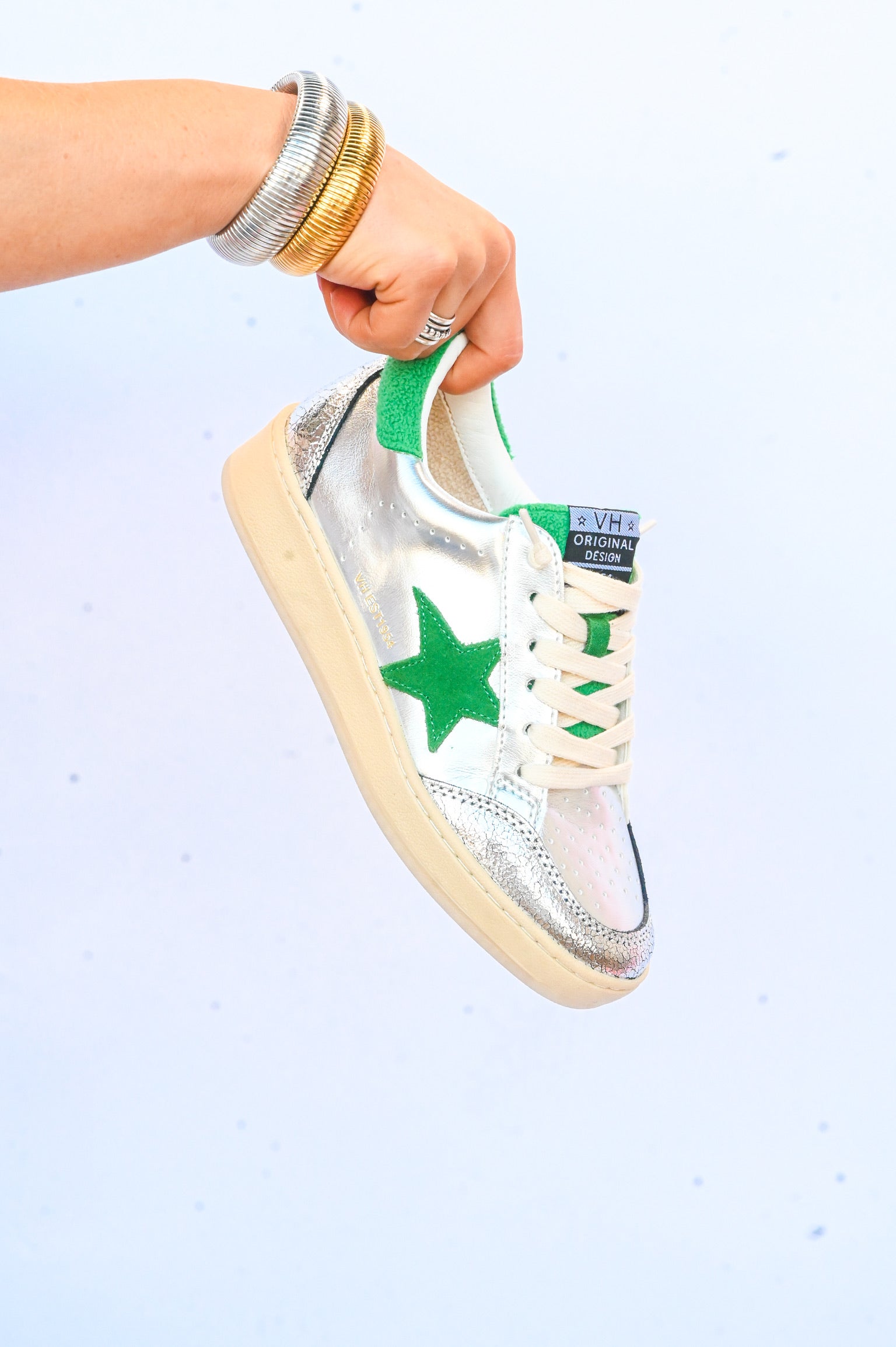 Hand holding metallic silver Vintage Havana sneakers with green star and white laces on white background